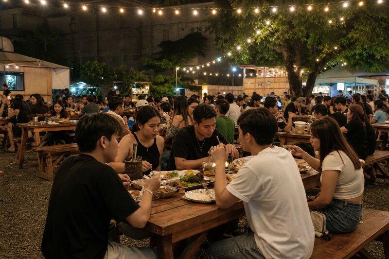 Communal wooden picnic tables filling an open-air food park at night, strung with warm fairy lights, groups of friends eating and talking