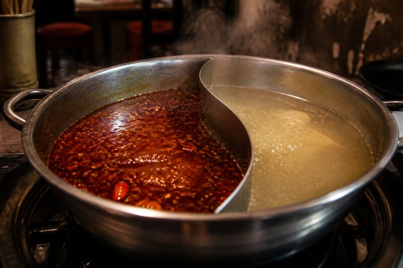A divided stainless-steel hotpot set into a restaurant table, one half filled with red chili-oil mala broth studded with peppercorns, the other a pale clear broth, steam rising