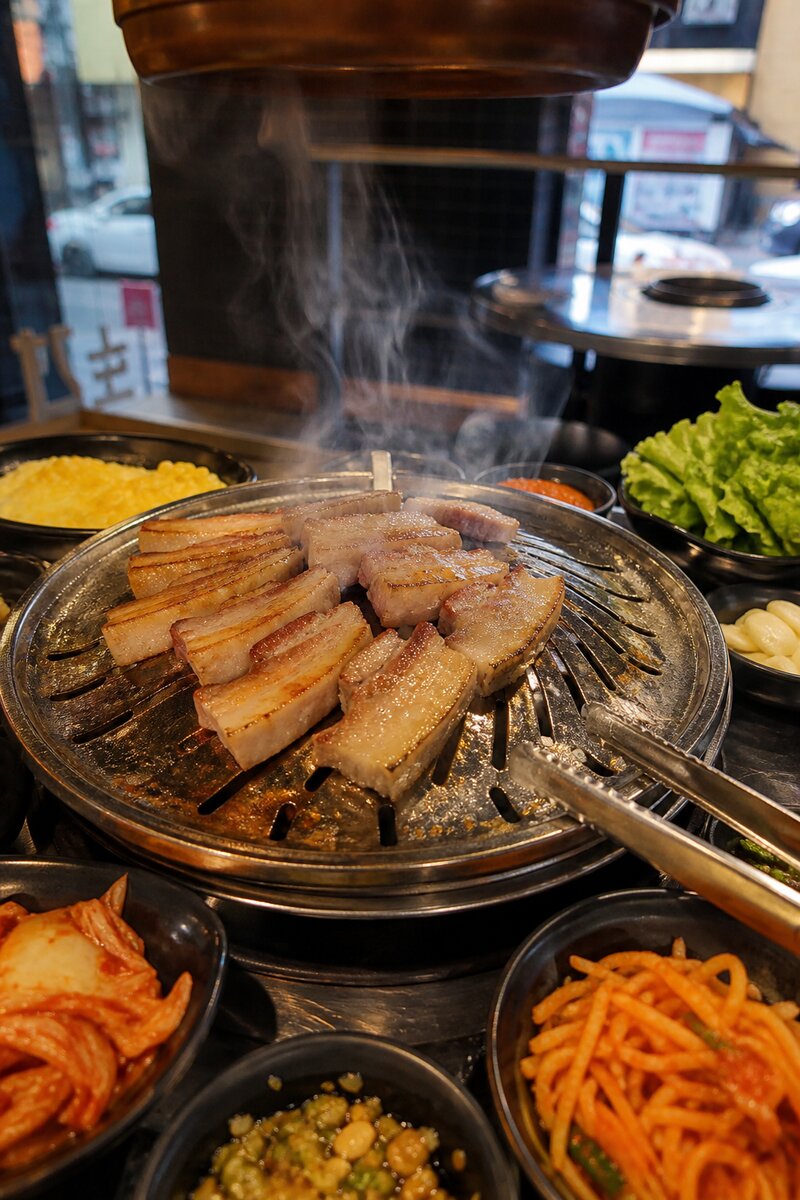 Thick slices of pork belly sizzling on a domed Korean BBQ grill at the center of a table, surrounded by colorful banchan side dishes