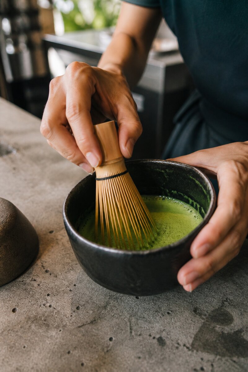 A barista whisking bright green ceremonial matcha in a bowl with a bamboo chasen at a slow bar