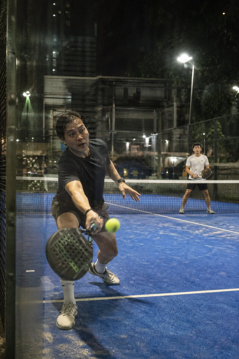 Two players mid-rally on a glass-walled padel court under bright floodlights