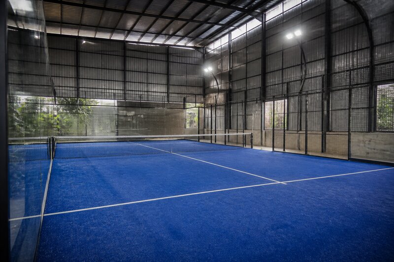 An empty indoor padel court with glass walls and blue turf under floodlights