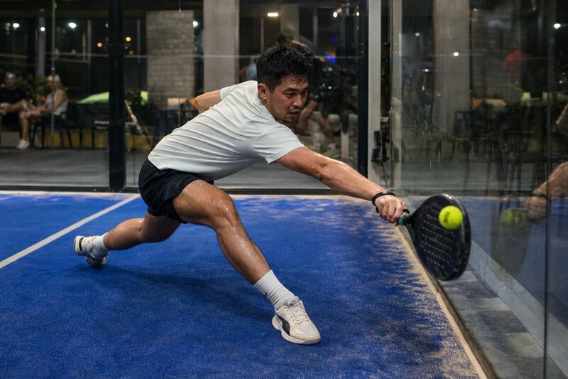 A player reaching to return the ball against the glass back wall of a padel court