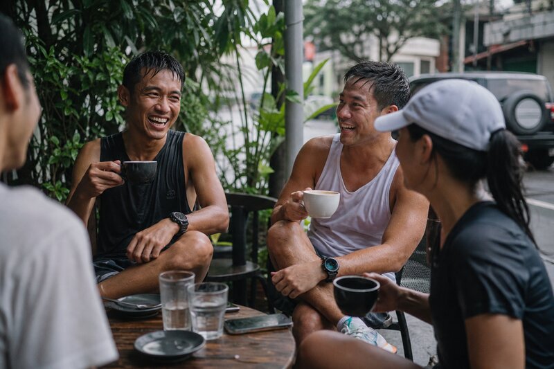 Runners gathered at an outdoor cafe with coffee cups after a morning run, laughing together