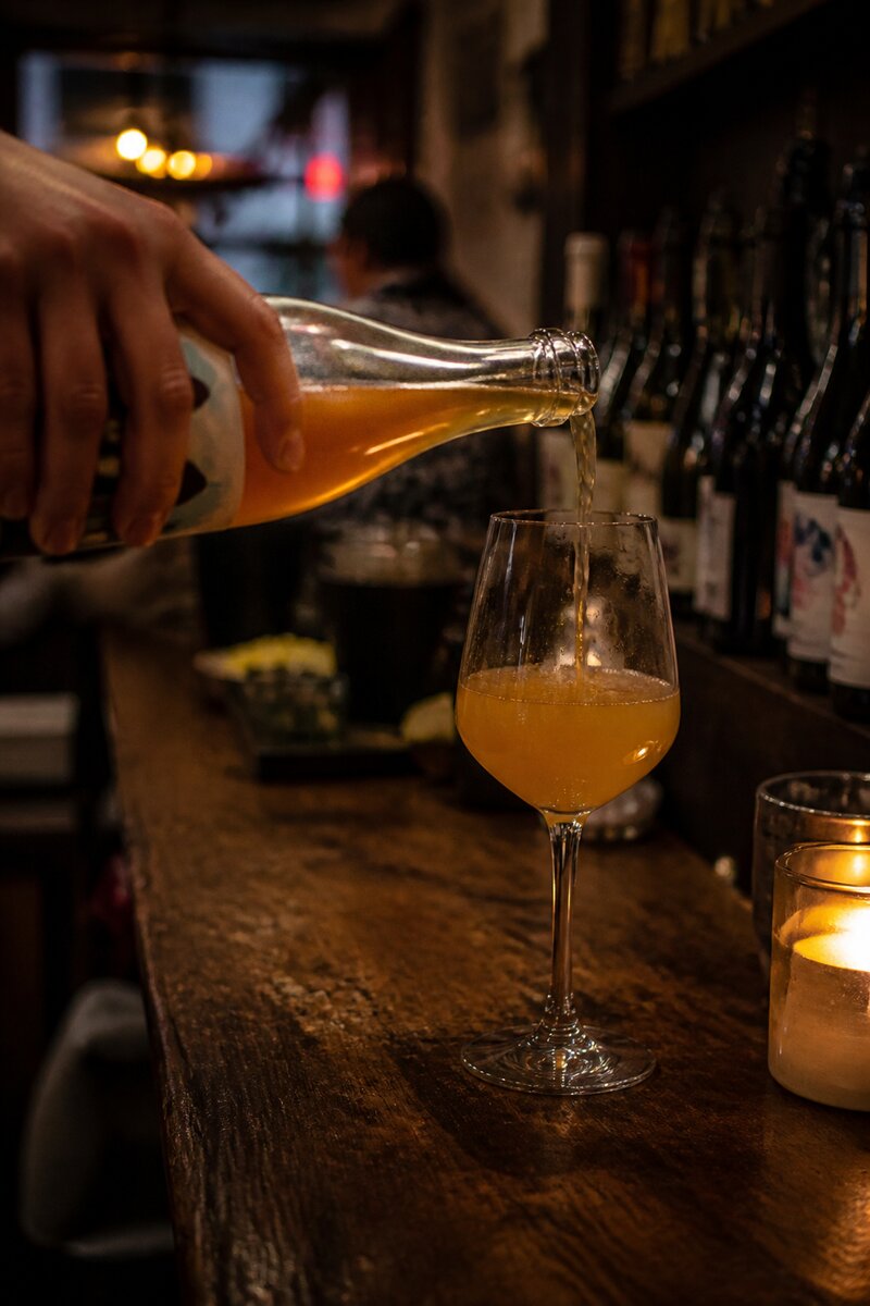 A hand pouring cloudy amber orange wine into a glass on a candlelit bar top, rows of natural wine bottles softly out of focus behind