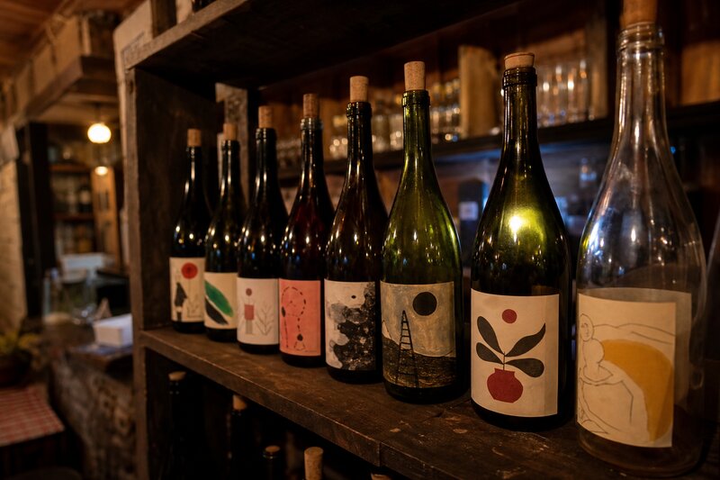 A shelf of natural wine bottles with hand-drawn artistic labels, softly lit in a cozy wine bar
