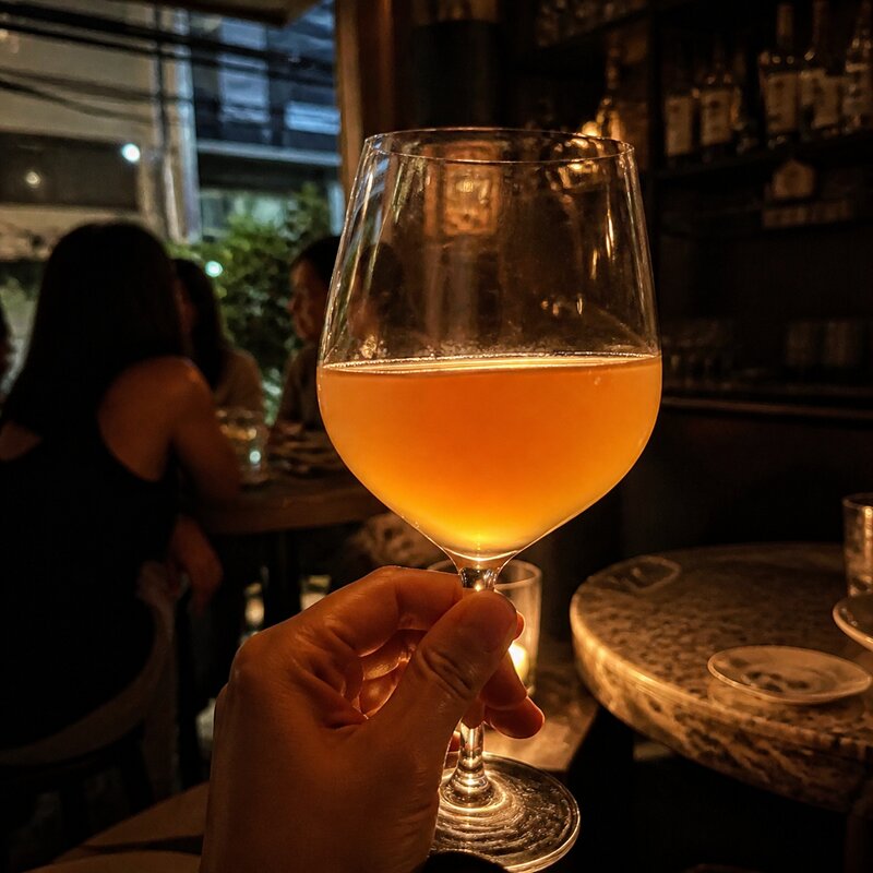 A glass of cloudy amber orange wine held up to warm light, showing its hazy, unfiltered color