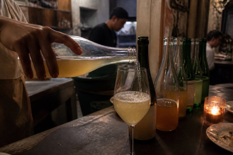 A close-up of cloudy pét-nat being poured into a wine glass at a dimly lit bar, bubbles rising, open bottles lined up on the counter