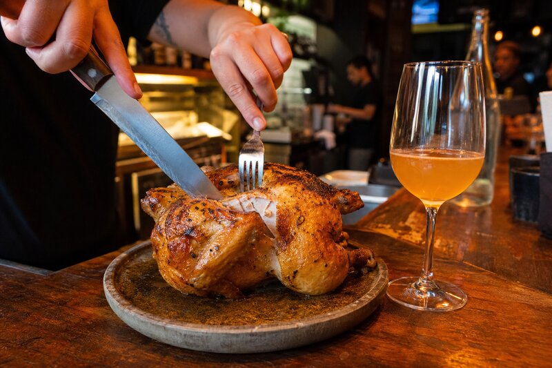 A golden koji-marinated rotisserie chicken being carved beside a glass of natural wine