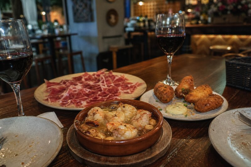 A shareable spread of Spanish tapas with glasses of red wine on a warm-lit bar table